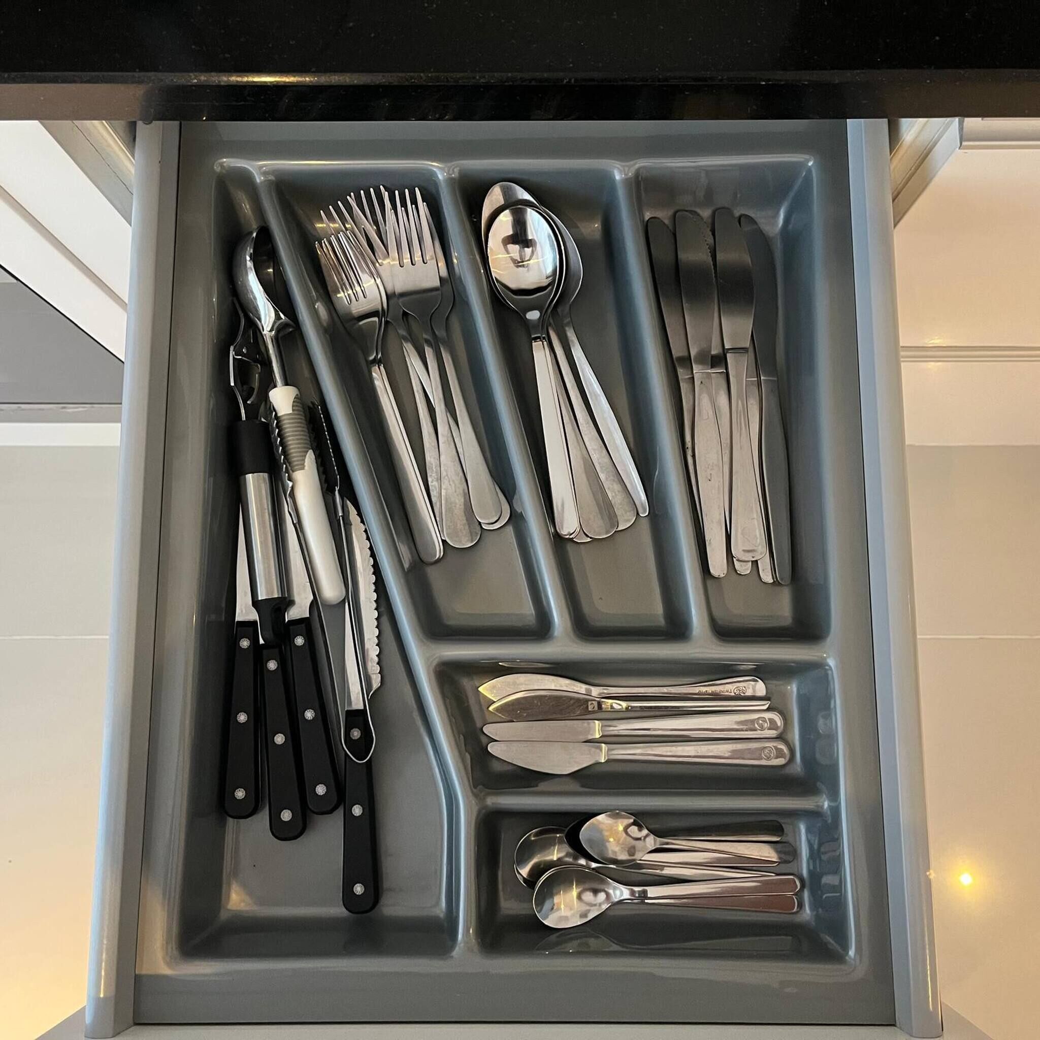 A drawer with compartments containing various kitchen utensils such as forks, knives, spoons, and a spoon rest.