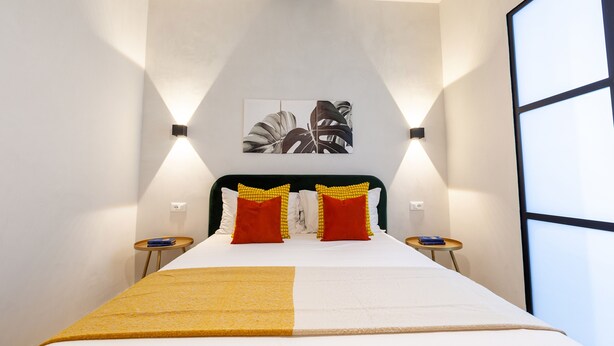 A neatly made bed with a yellow throw, two red pillows, and a black and white artwork above it. There are two bedside tables with lamps. - Malva Rooms (Agrigento)