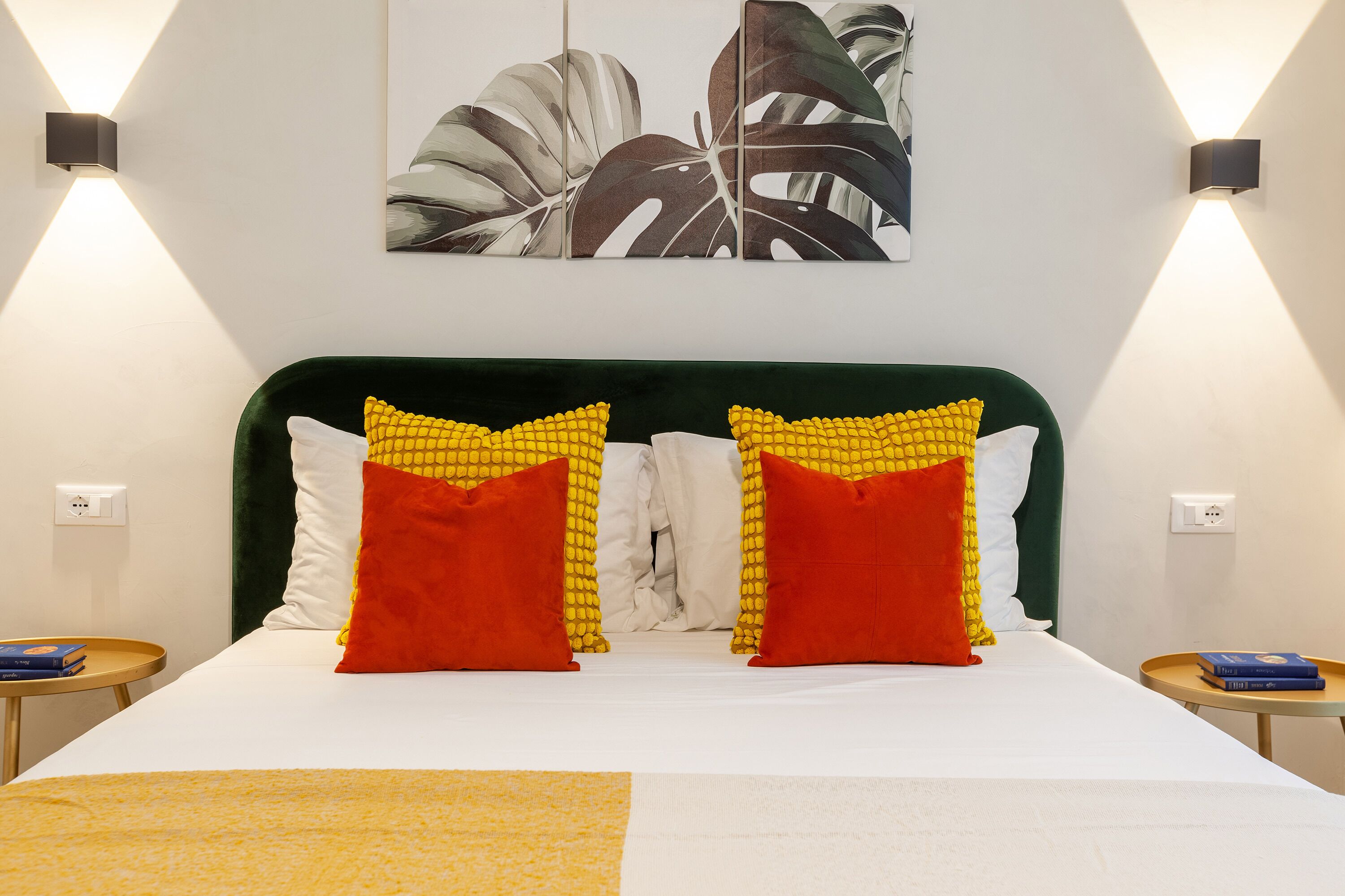 Una cama con cabecera tapizada en verde, dos almohadas con estampado amarillo y rojo, y una colcha blanca.