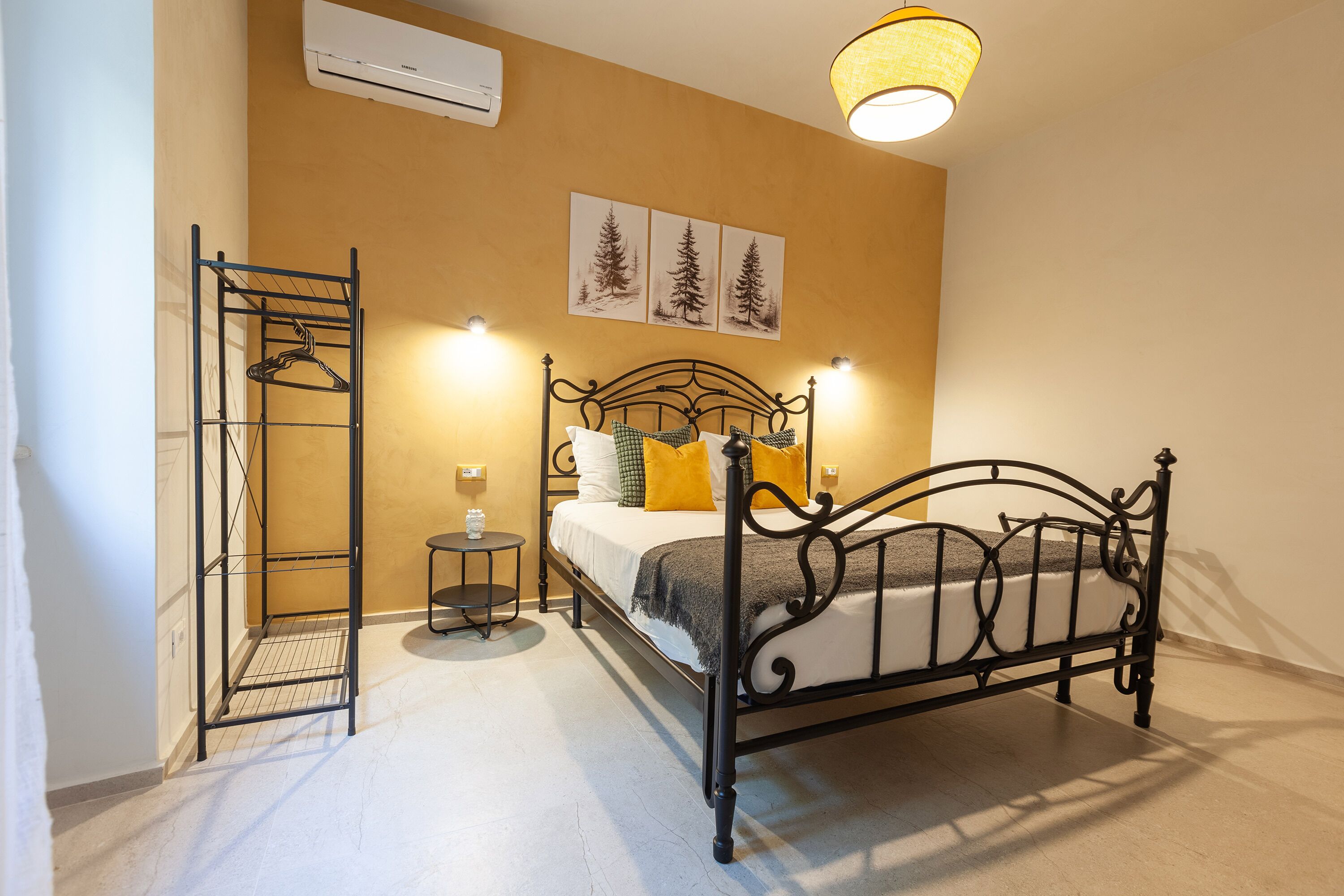 Un dormitorio con estructura metálica para la cama, una mesita de noche, un perchero y cuadros colgados en la pared.