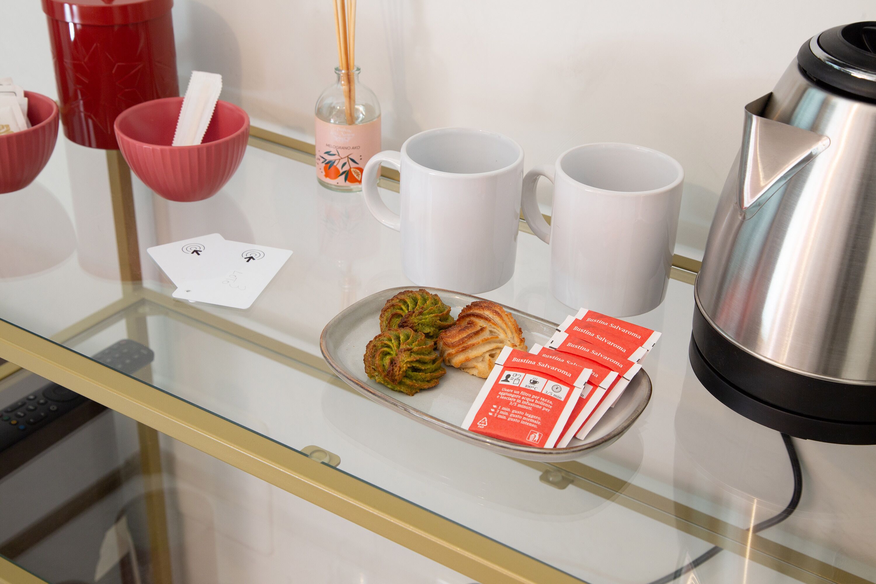 A glass table with a tray of pastries, two white mugs, a red mug, a glass bottle with a straw, and a stainless steel kettle.