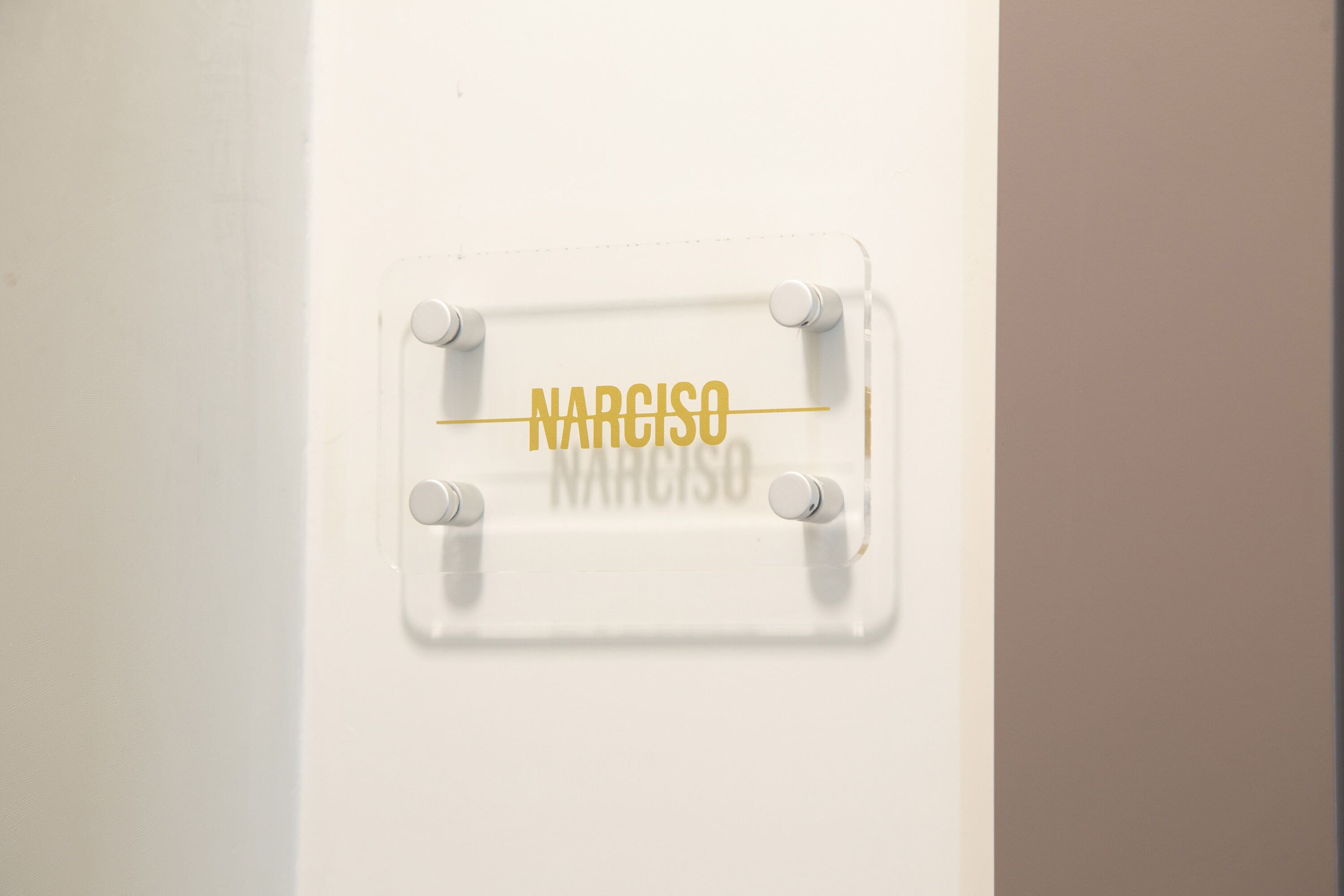 A white wall with a clear plastic sign that reads "NARCISO" in gold lettering.