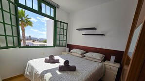 Quarto com cama, duas mesas de cabeceira, um criado-mudo, janela com moldura verde e vista para palmeiras e edifícios lá fora.