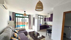 Sala de estar com sofá, mesa de centro e área de jantar com mesa e cadeiras. Há uma cozinha com geladeira e janela.