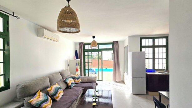 Sala de estar com sofá, mesa de centro, luminária pendente, área de cozinha com geladeira e vista para o exterior.