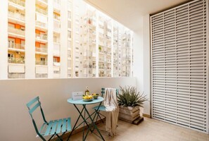 Un balcon donnant sur un immeuble à plusieurs étages, une table avec des chaises, une plante en pot et une fenêtre avec des stores.