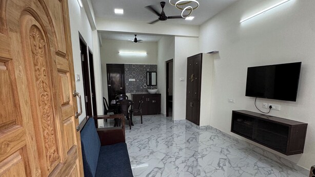 A spacious living area with a marble floor, a wooden door, a blue sofa, a ceiling fan, recessed lighting, and a flat-screen TV mounted on the wall.