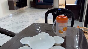 A dining table with white bowls and a water bottle, a sofa with a colorful cushion, a TV on the wall, and a yellow curtain.