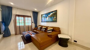 Uma sala de estar com um sofá de couro marrom, um banquinho azul com padrão da Union Jack, um quadro de paisagem emoldurado e uma lixeira preta com capa de renda.