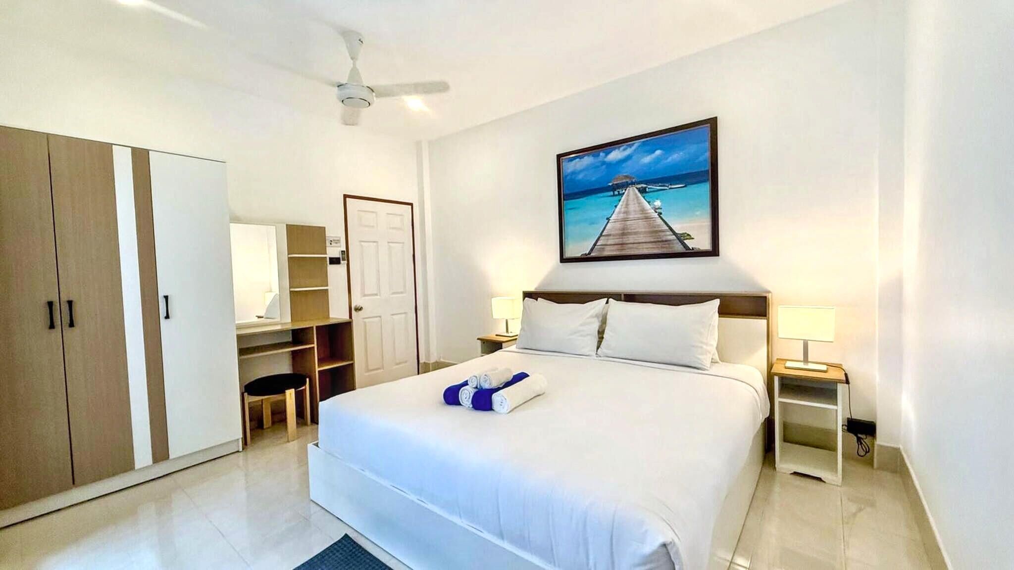 A hotel room with a bed, a ceiling fan, a painting of a pier, a wardrobe, a desk, and a chair.