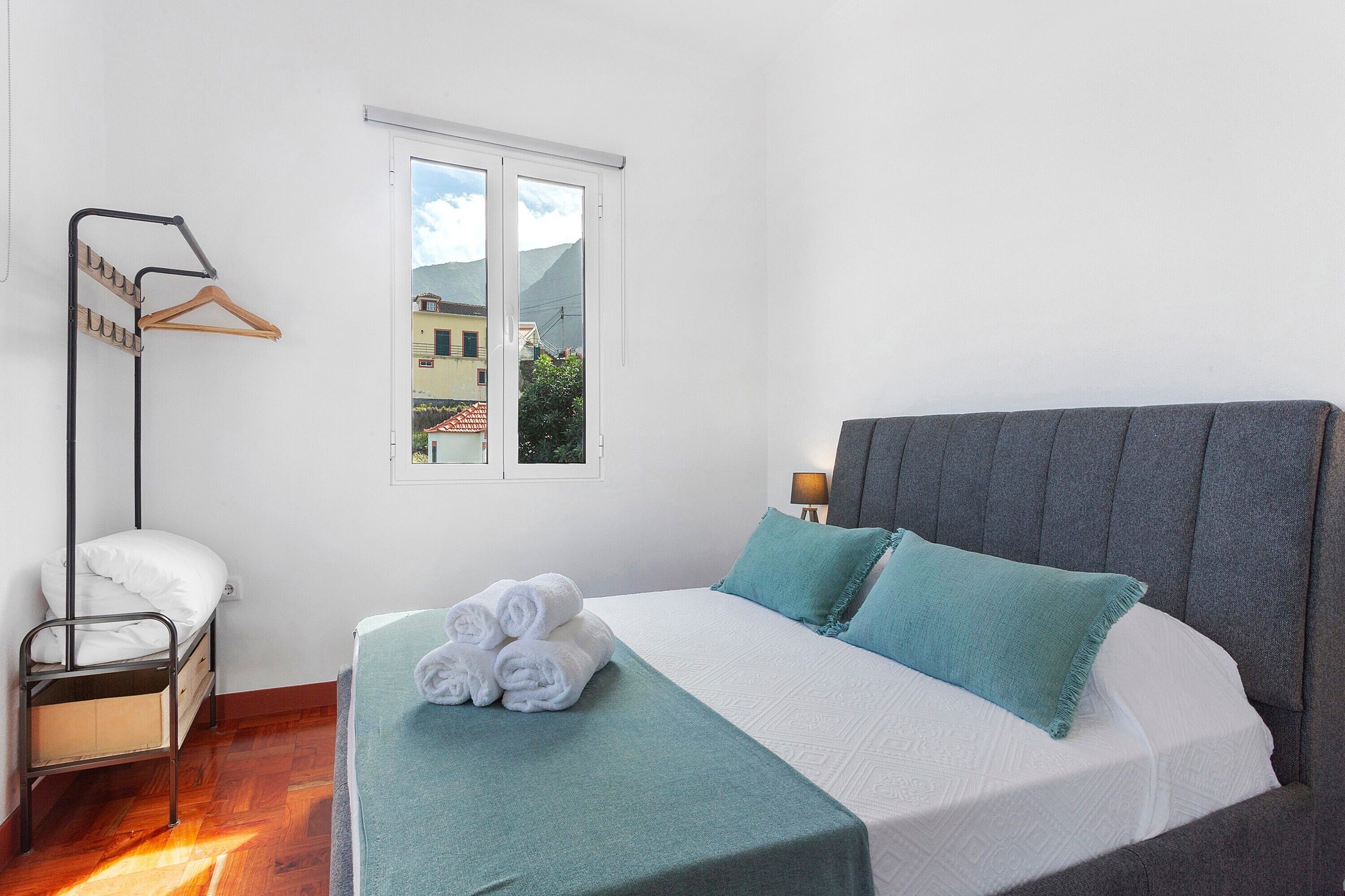 A single bed with a grey headboard, a bedside table with a lamp, a window with a view of buildings, and a metal rack with clothes.