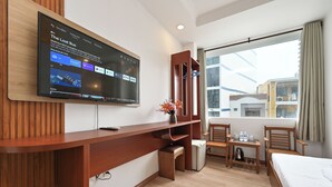 Une chambre d’hôtel équipée d’un téléviseur fixé au mur, d’un bureau en bois, d’une petite table avec des bouteilles d’eau, d’un lit et d’une fenêtre avec des rideaux.