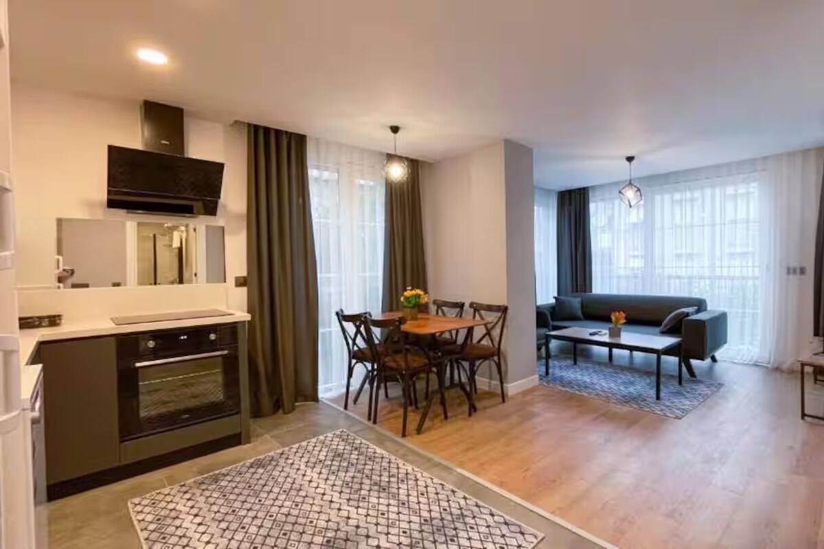 Una cucina moderna con forno integrato, un'area pranzo con tavolo e sedie, un divano e un'ampia finestra con tende.