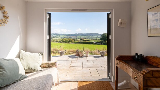 A room with a bed, a wooden desk, and a view of a green landscape through a glass door.