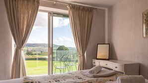 A bedroom with a large bed, a nightstand, a mirror, and a view of a green landscape through a sliding glass door.