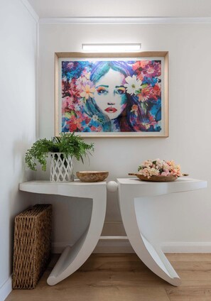 Une console blanche, avec un tableau encadré au-dessus, une plante en pot et un bol en bois posé sur la console.
