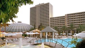 Zona de piscina d'un hotel amb un pont, gandules, ombrelles i un edifici gran al fons.