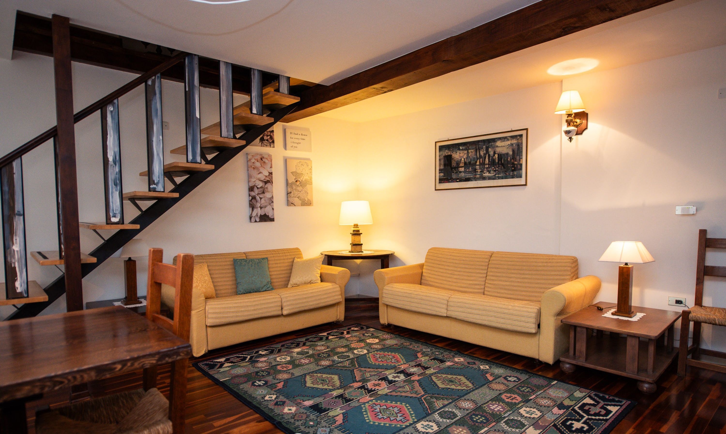 A cozy living room with two sofas, a patterned rug, wooden flooring, a staircase, and wall-mounted lamps.