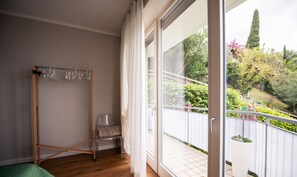 Una stanza con pavimento in legno, un letto verde, un armadio in legno e una porta in vetro che dà su un balcone con recinzione bianca e vegetazione.