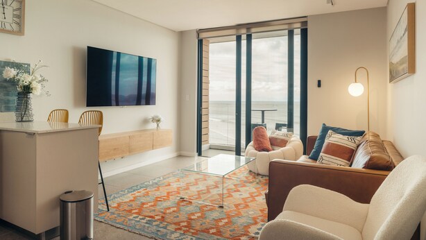 Una sala de estar moderna con un televisor grande, un sofá, una mesa de centro y vistas al océano a través de amplios ventanales.