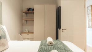 Una habitación de hotel con una cama, un edredón verde con textura, una toalla enrollada, un armario, un estante con libros y artículos de aseo, y una puerta con espejo.