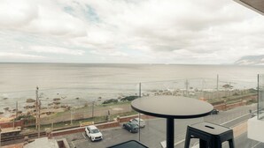 Un balcón con mesa y sillas que da a una carretera costera con coches aparcados y vistas al mar.