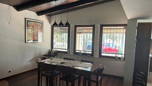 A dining area with a wooden table and chairs, large windows with metal bars, and a ceiling with exposed beams.