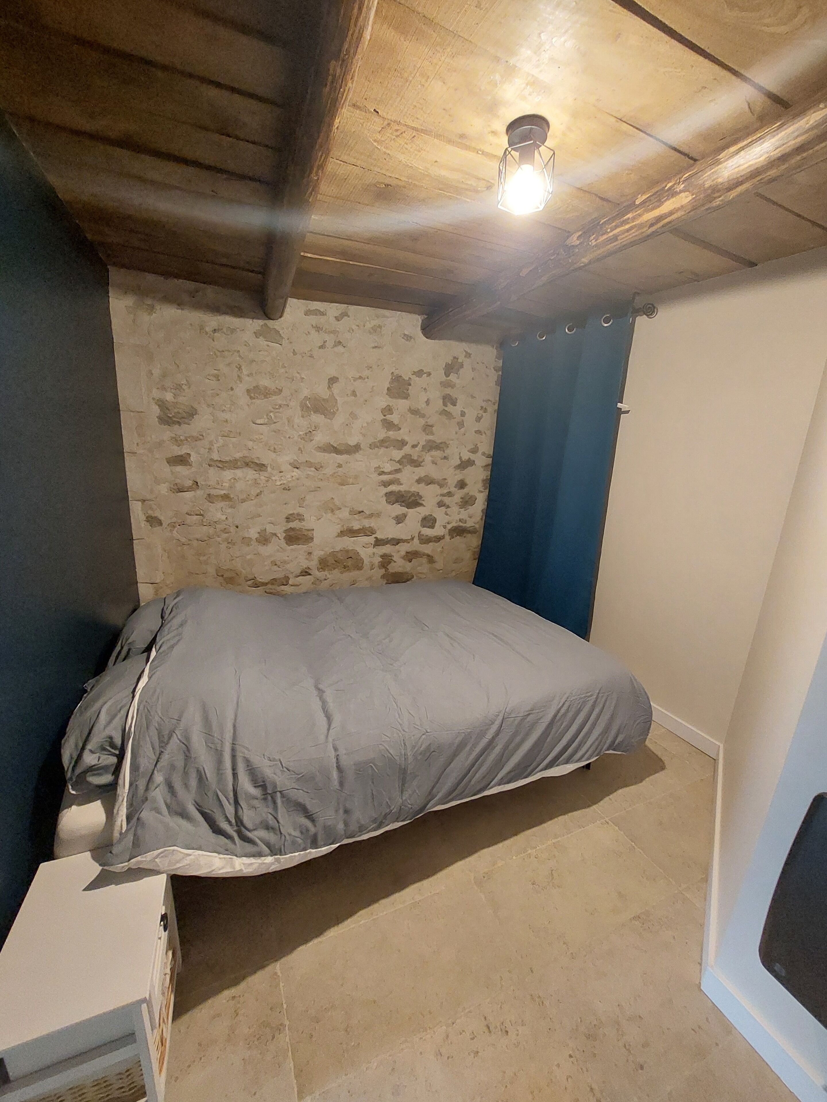 Una habitación pequeña y rústica con una cama individual, un muro de piedra y techo de madera.