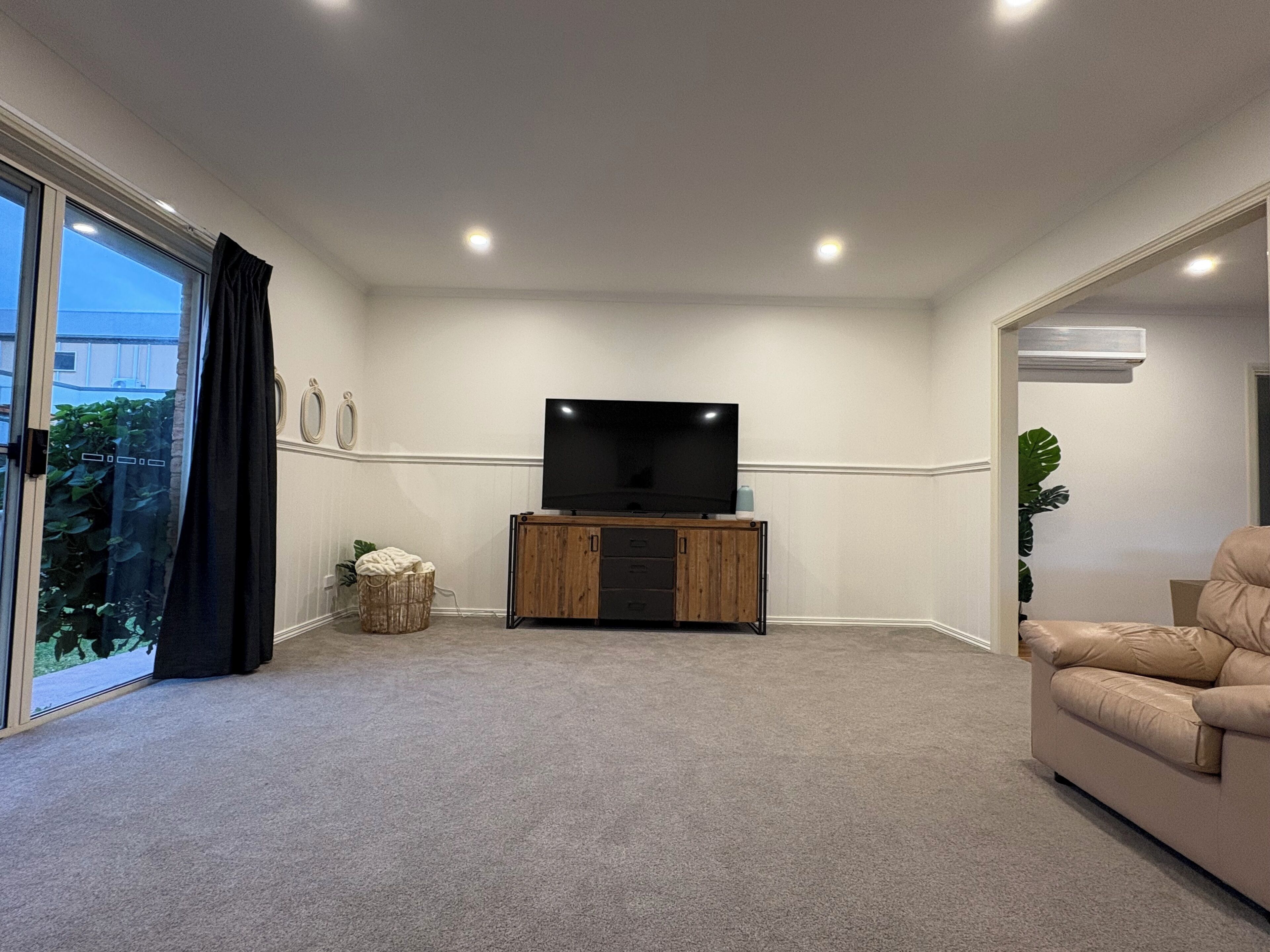 A living room with a flat-screen TV on a wooden stand, carpeted floor, and a beige sofa.