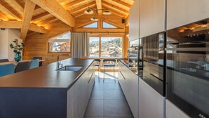 A modern kitchen with wooden beams, a large island, and a view of mountains. - Hameau des clos Téléporté (Les Gets)