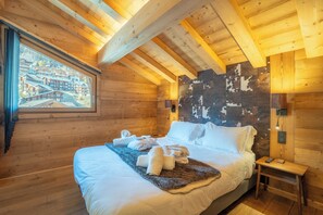 A wooden cabin room with a bed, towels, a small table, and a painting of a village. - Hameau des clos Téléporté (Les Gets)