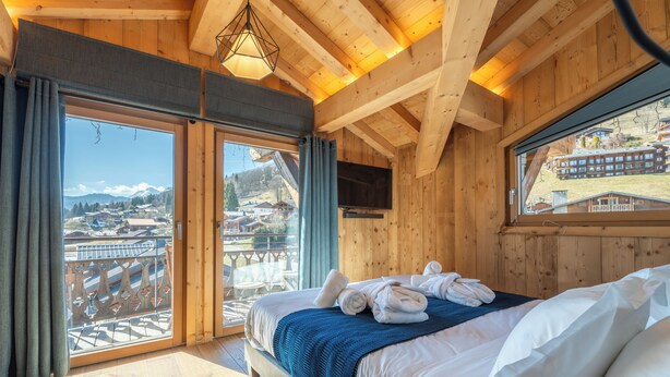 A wooden cabin room with a bed, a TV, and a view of a mountain village. - Hameau des clos Téléporté (Les Gets)