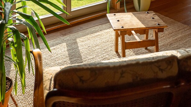 Une pièce chaleureuse avec un plafond en bois, une grande porte-fenêtre coulissante donnant sur une cour verdoyante, une petite table en bois et une chaise.