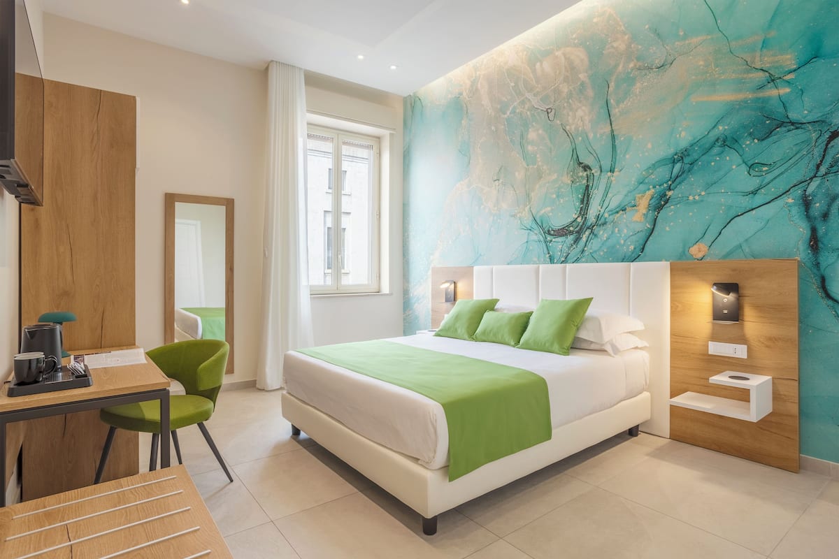 Ein modernes Hotelzimmer mit einem großen Bett, grünen Kissen, einem Schreibtisch mit Stuhl, einem Holzschrank und einem großen, abstrakten Wandbild.