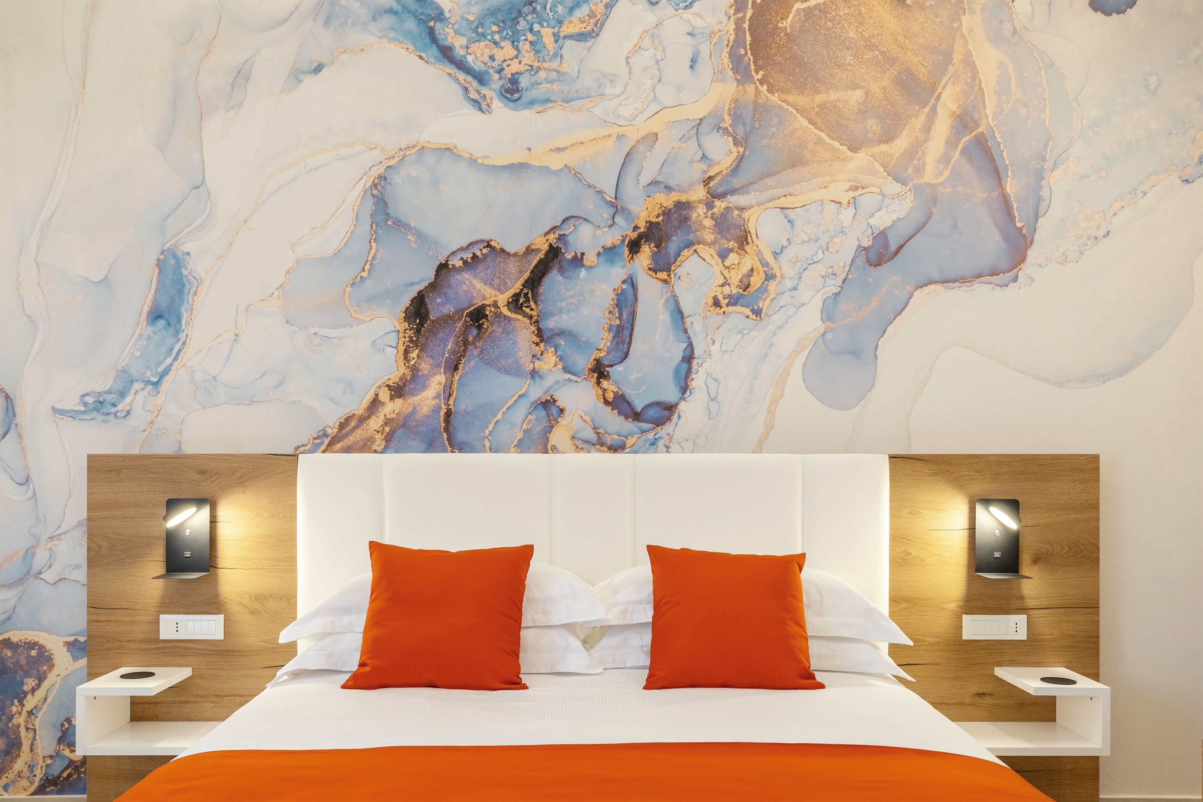 A hotel room with a marble wall, a bed with white and orange bedding, and bedside tables with lamps.
