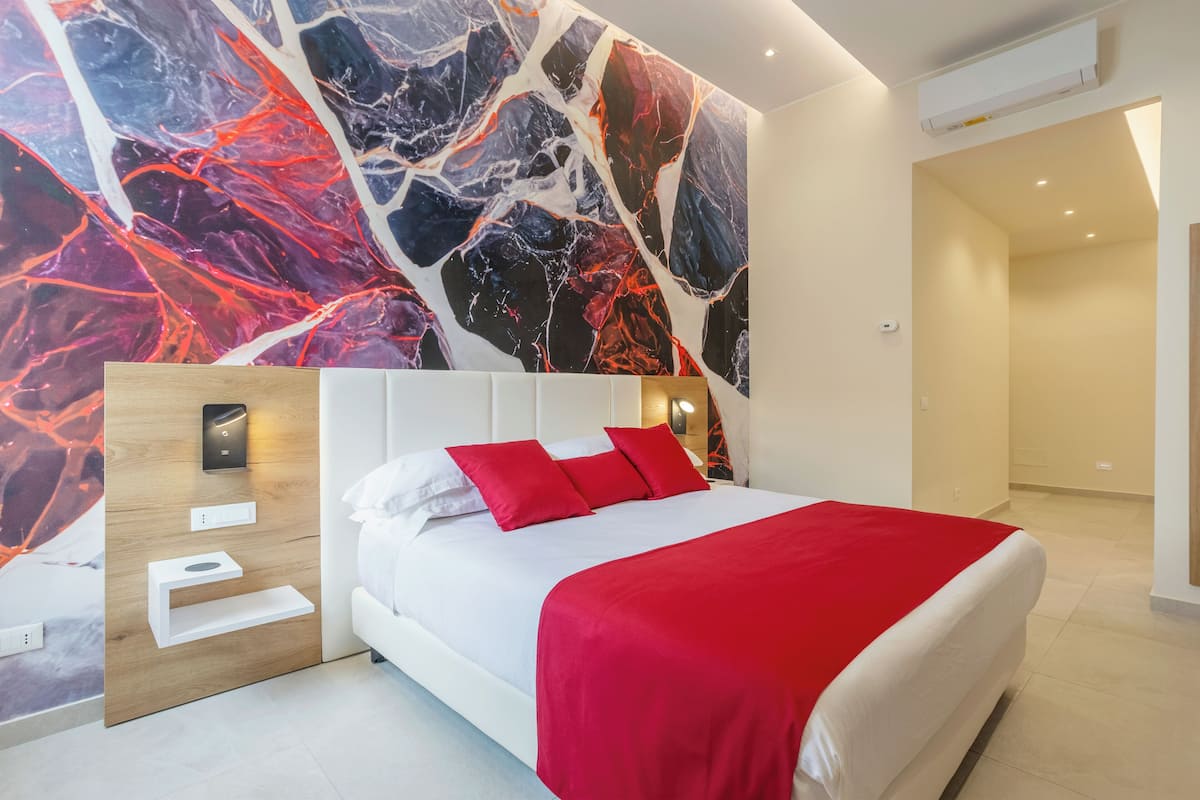 Ein modernes Schlafzimmer mit einem weißen Bett, roten Kissen und einer roten Decke. Darüber hängt eine Wanddekoration aus Marmor, und an der Wand befindet sich ein Holzpaneel mit einer Leuchte.