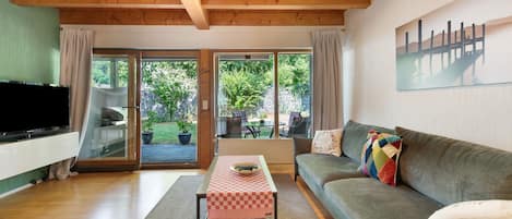 Ein Wohnzimmer mit Holzboden, einer Couch, einem Fernseher und Blick auf einen Garten durch Glasüren.