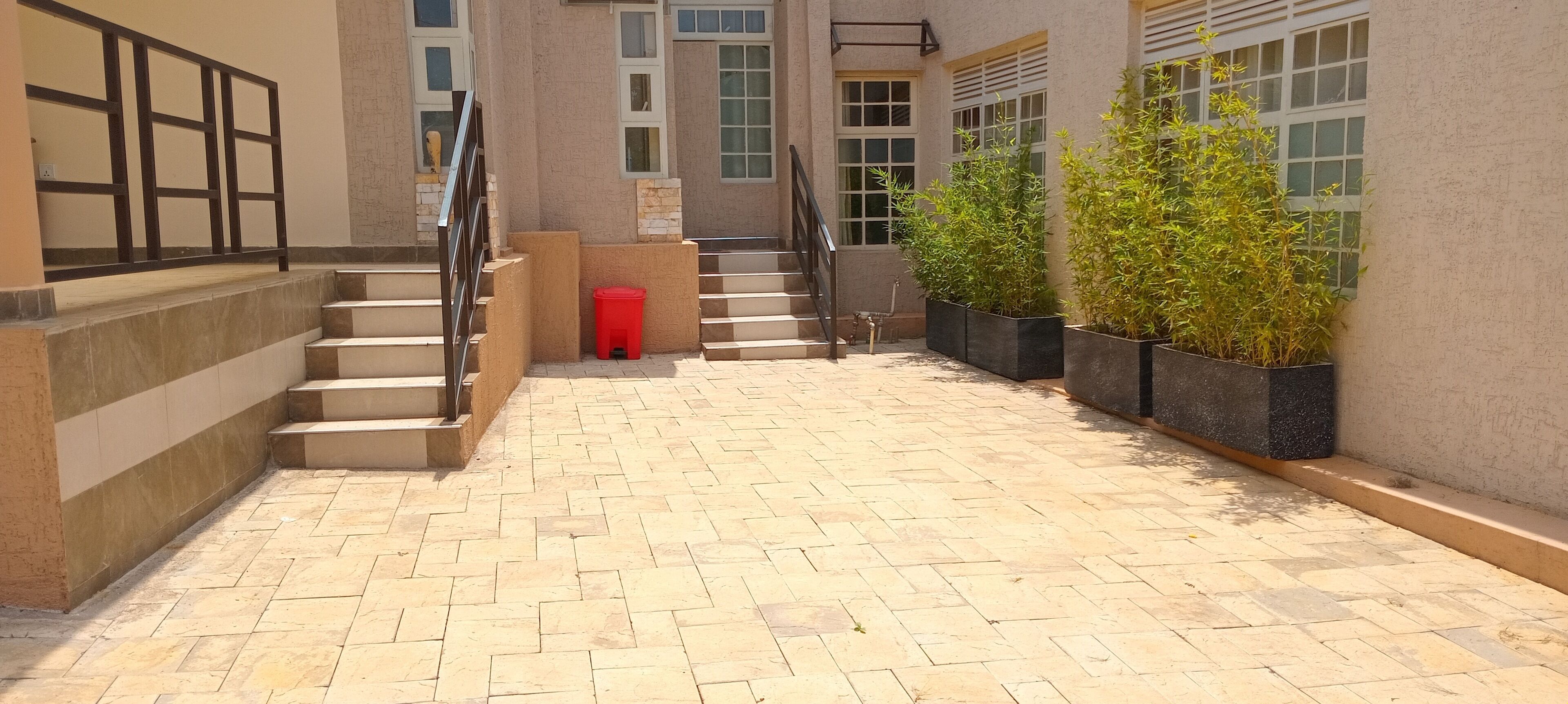 Un patio empedrado con una escalera, plantas en macetas y un bote de basura rojo.