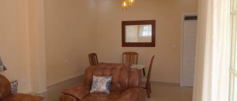 A living room with a brown leather sofa, a wooden dining table with chairs, a mirror on the wall, and a white door in the background.