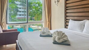 A hotel room with a large bed, two towels, a window with a view of palm trees and a pool, and a wooden headboard. - Haven Lagoon Condominium (Patong)