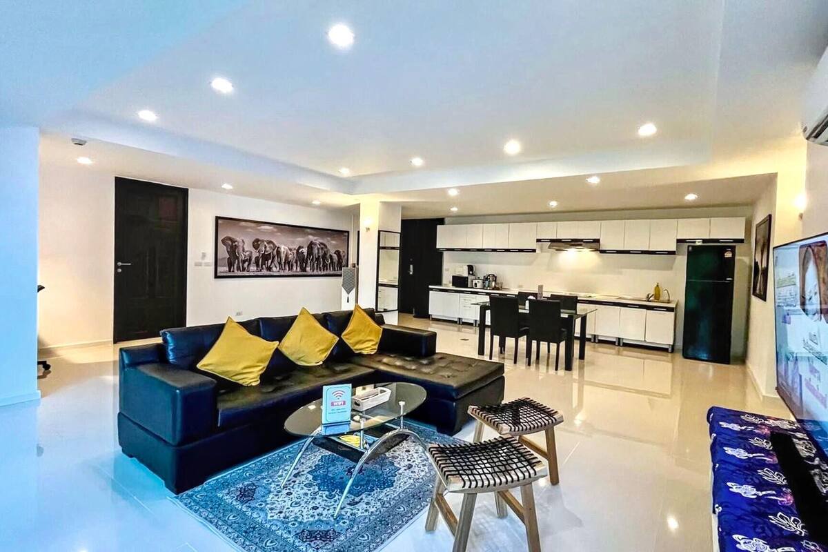 A modern living room with a sofa, coffee table, and chairs, a kitchen with white cabinets, and a dining area.