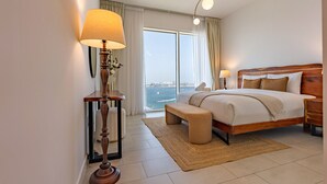 A hotel room with a large bed, bedside lamps, a wooden headboard, a bench, a view of the sea, and a large window.