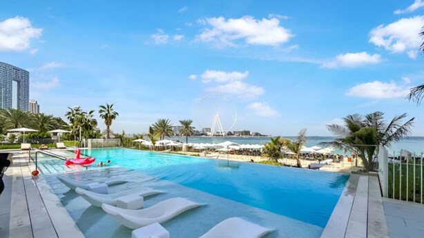 A swimming pool with lounge chairs, a Ferris wheel, and a beach in the background.