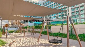 A playground with swings and a slide, surrounded by sand and greenery, with a modern building in the background.