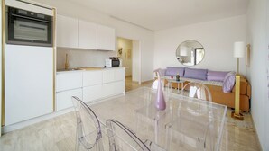 Una cocina moderna con armarios blancos, horno empotrado y microondas. Se observa un comedor con una mesa de vidrio transparente y sillas. Al fondo, un sofá con cojines.