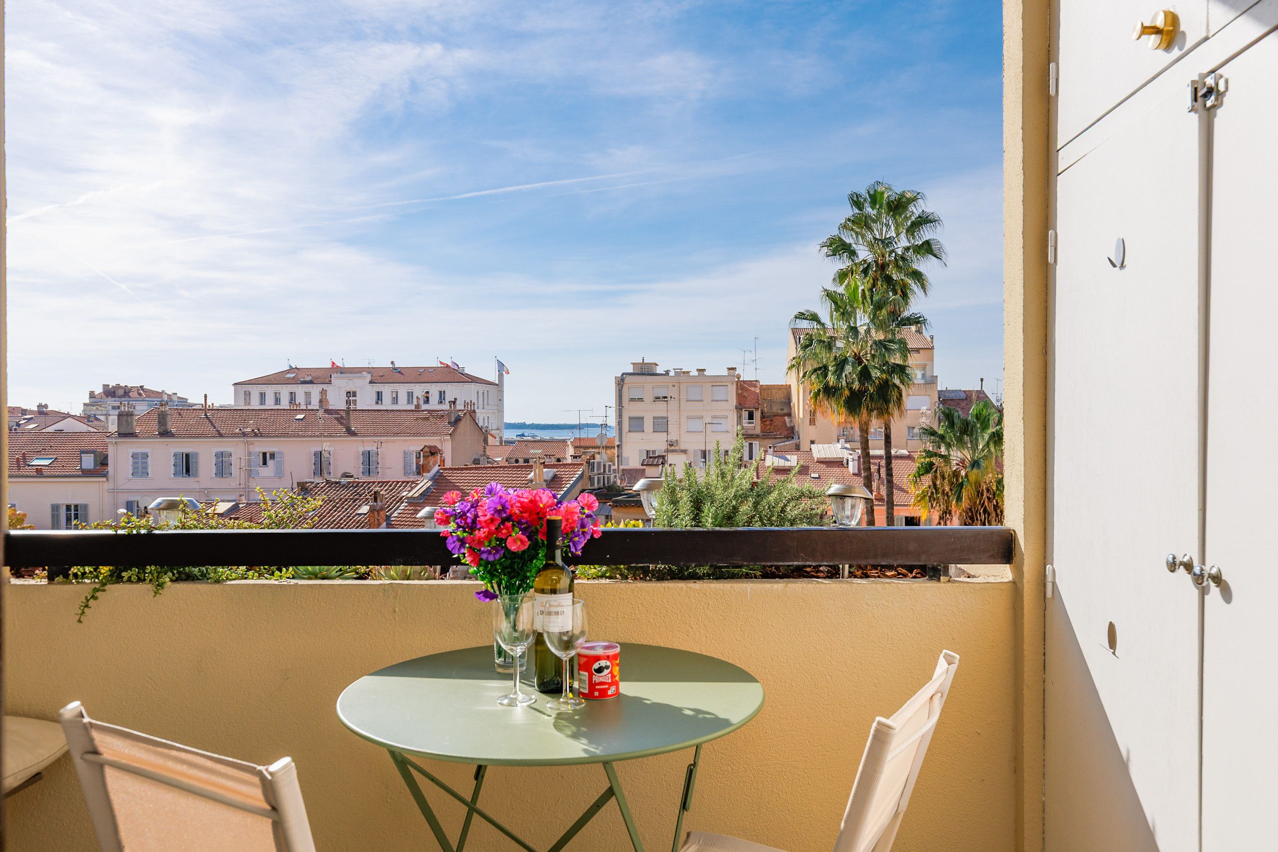 A balcony with a table, chairs, a vase of flowers, a wine bottle, and a can of Pringles.