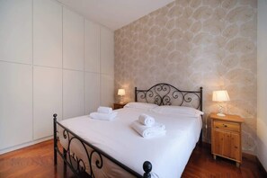 Cama bem arrumada com roupa de cama branca e toalhas dobradas, mesa de cabeceira de madeira com abajur e papel de parede estampado na parede.
