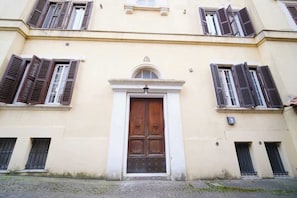 Un bâtiment doté d’une porte centrale en bois, de nombreuses fenêtres avec volets et d’un sol pavé.