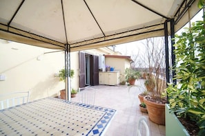 A covered patio with a table and chairs, potted plants, and a view of a building with a door and windows.