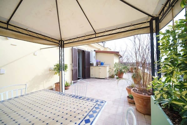 Une terrasse couverte avec une table et des chaises, des plantes en pot et une vue sur un bâtiment doté d’une porte et de fenêtres.